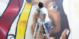 Artista no alto da escada grafitando muro. Muro parcialmente pintado, exibe rosto e faixas coloridas