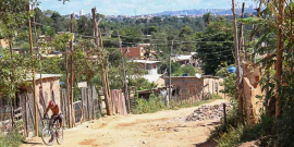 Estrada de terra com material de construção à direita e homem de bicicleta à esquerda. Nos dois lados, barrações com tijolos aparentes, durante o dia. 