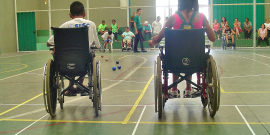 Dois estudantes cadeirantes, de costas, em quadra esportiva, durante o dia; no meio, cinco bolas pequenas no chão. Ao fundo, três estudantes cadeirantes e um homem de pé. Mais ao fundo, atrás de uma grade de proteção, cerca de dez adultos acompanham sentandos em arquibancada. 