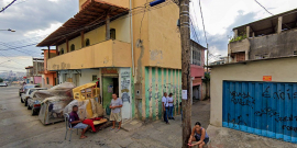 Beco à direita em rua com alguns carros cobertos ao fundo e casa com muro pixado à direita, durante o dia