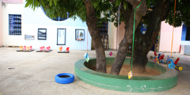 Pátio de escola infantil com brinquedos ao fundo e  vários livros pendurados em uma árvore robusta.