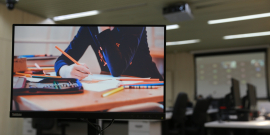 tela de computador mostra aluno sentado estudando; ao fundo, mobiliário do plenário 