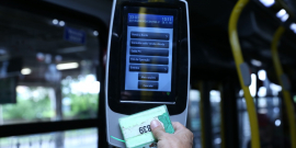 Mão humana passa cartão em máquina de bilhetagem eletrônica, dentro de ônibus, à noite.