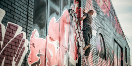 Homem em cima de escada, pinta muro urbano com desenhjos coloridos, durante o dia