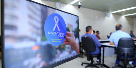 Em tela, laço branco em fundo azul, com os dizeres Novembro Azul. Ao fundo, imagem de homens que participam da palestra