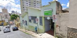 Fachada de Centro de Saúde Horto, com cidadã caminhando em frente, na calçada.  a rua é inclinada, e mais de cinco carros aparecem no final da imagem diurna, em direção contrária à do centro de saúde. 