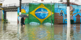 Imagem de uma pessoa atravessando com um  guarda-chuva, uma área totalmente alagada. Ao fundo, um muro com a bandeira do Brasil pintada. 