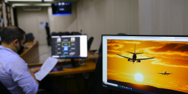 Tela mostra avião sobrevoando a cidade com um céu alaranjado, indicando o pôr-do-sol. Um homem caucasiano , vestido com uma camisa azul de mangas compridas, está sentado ao lado segurando algumas folhas de papel , 