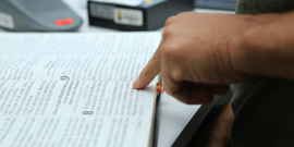 Um livro aberto e uma pessoa acompanha as letras com o dedo indicador fazendo inferência à uma leitura.