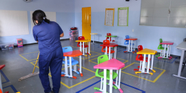 Mulher de conjunto de calça e blusa azul e máscara, de costas, limpa sala de Unidade de Educação Infantil com rodo. 