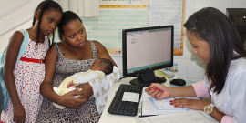 Mulher sentada com filho recém nascido no colo e filha de pé, ao lado, é atendida por médica que preenche um formulário, sentada à frete de mesa com computador. 