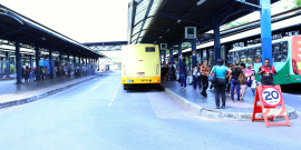 Na Estação Diamante  passageiros, em pé, aguardam o ônibus. Estacionado, um ônibus amarelo aguarda a saída