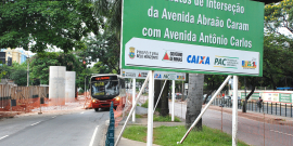 Placa indicativa de obras na Avenida Abraão Caram com Avenida Antônio Carlos. Trânsito parcialmente fechado com manilhas. Um ônibus passa no estreitamento da pista 