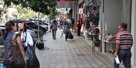 Calçada ocupada por pedestres fazendo compras em lojas diante de um estacionamento repleto de carros. Duas mulheres cheias de sacola conversam paradas na calçada