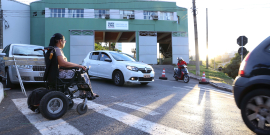 Cadeirante atravessa a rua em frente à Polícia Militar na faixa de pedestres, durante o dia. À esquerda, carro parado espera a travessia. 
