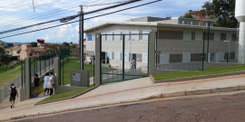 Fachada do Centro de Saúde Havaí, uma casa de dois andares com duas portas e seis janelas frontais, gramas so redor e quatro pessoas atravessando a porta da grade de acesso, durante o dia. 