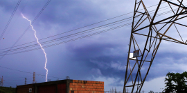 Imagem de uma torre de celular ao lado de uma edificação com tijolos à mostra . Ao fundo , um céu nublado cortado por um raio
