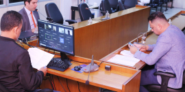 Três vereadores sentados à mesa em reunião presencial, um deles observa tela de computador com participantes da reunião de maneira remota. 