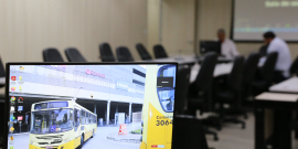 Imagem na tela do computador mostra ônibus da linha 328, amarelo, contornando um canteiro central em frente a um grande estabelecimento comercial . ao fundo, imagem desfocada de dois vereadores sentados à mesa