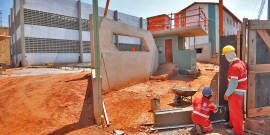 Dois homens de capacete e uniforme laranja da Prefeitura de Belo Horizone, trabalham em obra, erguendo parede