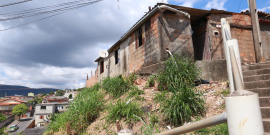 Casebre com tijolos aparentes e telhado de zinco no alto de um barranco com mato alto