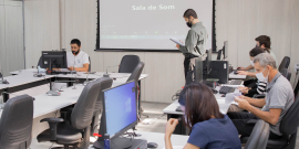 Vereador sentado à mesa, preside reunião semipresencial. Em plenário, cinco servidores da Câmara auxiliam na reunião