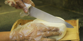 Queijo branco em tábua de madeira sendo cortado por mãos enluvadas.