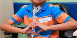 Criança vestida com camisa listrada de azul e laranja, com um aparelho ligado na traqueia, sentado em uma cadeira. Ele faz um gesto de coração com as duas mãos à frente do corpo