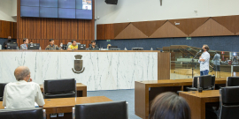 Quatro homens sentados à mesa do Plenário Amynthas de Barros e um homem falando ao microfone, à direita. Ao fundo, quatro assessores da Câmara.
