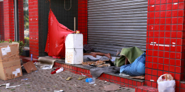 Pessoa em situação de rua , deitado na porta de um estabelecimento comercial fechado. Um pano vermelho está estendido cobrindo uma parte da calçada e há muito lixo na calçada