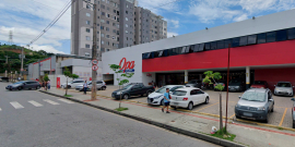 Rua em frente a supermercado, com carros estacionados e faixa para pedestres, durante o dia. 