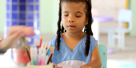Aluna com deficiência visualpassa suas mãos em caderno em espiral, sentada em carteira escolar. À sua frente, desfocado, um porta lápis com lápis coloridos e mãos femininas. 