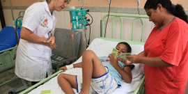 Criança em leito hospitalar acompanhada de mulher e atendida por profissional de saúde.