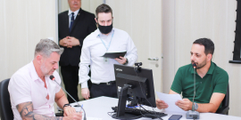 Dois vereadores em reunião presencial sentados à mesa, um deles em frente ao computador. Ao fundo, de pé, dois assessores da Câmara.