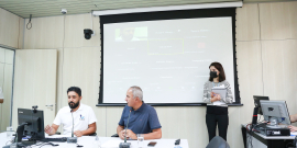 Os vereadores José Ferreira e Claudio do Mundo Novo estão sentado à mesa de reunião. Ao fundo e no alto, o telão mostra a imagem dos outros participantes da reunião. Em pé, uma mulher  usando uma blusa listrada, calça preta e máscara, segura uma pasta de documentos