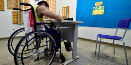 Criança em cadeira de Rodas escreve em carteira escolar, em sala de aula, em frente a um quadro azul.