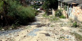 Imagem de uma rua íngreme de terra com mato alto de um lado e casas sem reboco de outro.