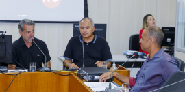 Imagem dos vereadores Wilsinho da Tabu, Claudio do Mundo Novo e Wagner Ferreira sentados em volta da mesa em U, durante a reunião. Ao fundo o telão identifica a participação remota do vereador Rubão. 