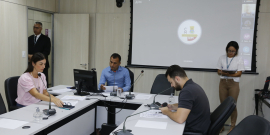 Três membros da Comissão em reunião, em torno de uma mesa em formato de U. Na imagem é possível ver um segurança na porta e uma servidora de pé auxiliando na reunião