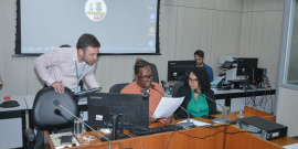 Dois parlamentares reunidos, sentados à mesa, auxiliados por um assessor técnico da câmara, de pé, ao lado deles. 