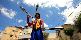 Homem com pernas de pau e vestido de palhaço faz malabares em dia ensolarado.
