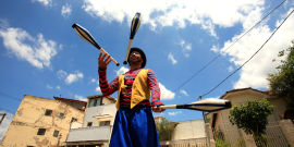 Homem com pernas de pau e vestido de palhaço faz malabares em dia ensolarado.