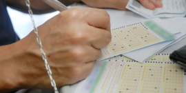 Mãos humanas preenchem boletos de loteria com números em sequência, em cima de um balcão. 