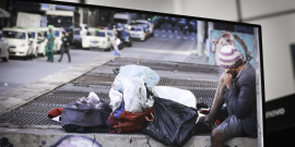 Pessoa em situação de rua sentada na calaçada diante de seus pertences. O homem usa uma blusa de brio, touca e bermuda