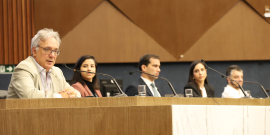 TRês homens e duas mulheres sentados à mesa. O primeiro deles, à esquerda, palestra.