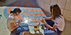 Duas garotas, sentadas, completam mosaico de cerâmica no chão à mão, durante o dia