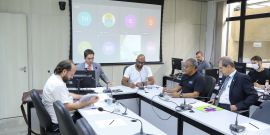 Cinco homens sentados à mesa, em reunião presencial.