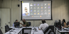 Três vereadores participam de reunião, sentados à mesa. No telão, é exibida reunião vistual com mais de 20 participantes, entre eles o secretário municipal de Planejamento, Orçamento e Gestão, André Reis.