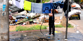 Imagem de uma pessoa tomando banho de mangueira. ela está atrás de um varal cheiro de roupas que secam ao ar livre, em plena praça