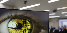 Imagem de um olho verde na tela do computado. É possível identificar uma menina de cabeça baixa refletida na imagem